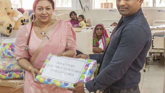 A member of the Colombo Court team handing a wrapped parcel of bed linen to a doctor in a hospital ward, children in the beds behind.