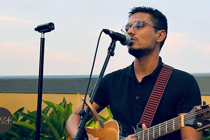 A musician singing into a microphone with an acoustic guitar on the rooftop at dusk, the sea behind him.