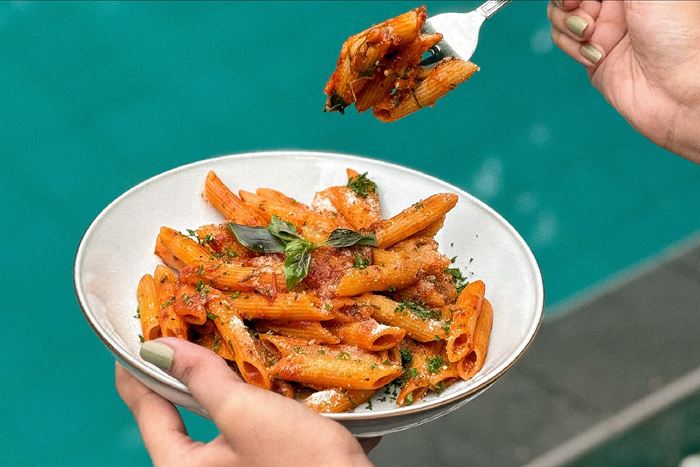 A plate of penne in tomato sauce held over the turquoise water of the pool at Colombo Court.