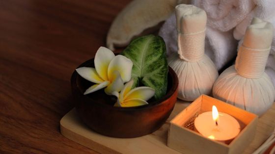 A spa tray with frangipani flowers, herbal compress balls and a lit candle on a timber board.
