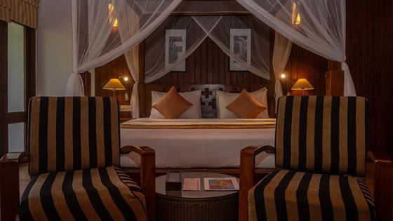 A suite at Colombo Court at night, a four-poster bed under white drapes with striped armchairs at its foot, warmly lit.