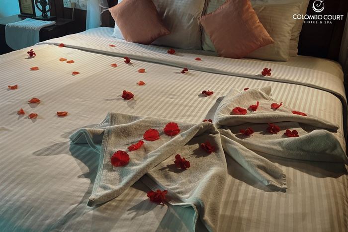 A large flower folded from towels laid across a white bed and scattered with red rose petals.