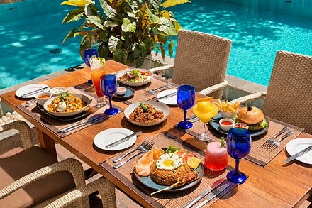 A shared lunch across a timber table at Amber Poolside, several plates and drinks beside the pool.