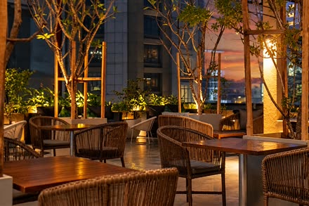 Tables and cane chairs on the rooftop at Cloud Café & Bar at twilight, trees lit from below.