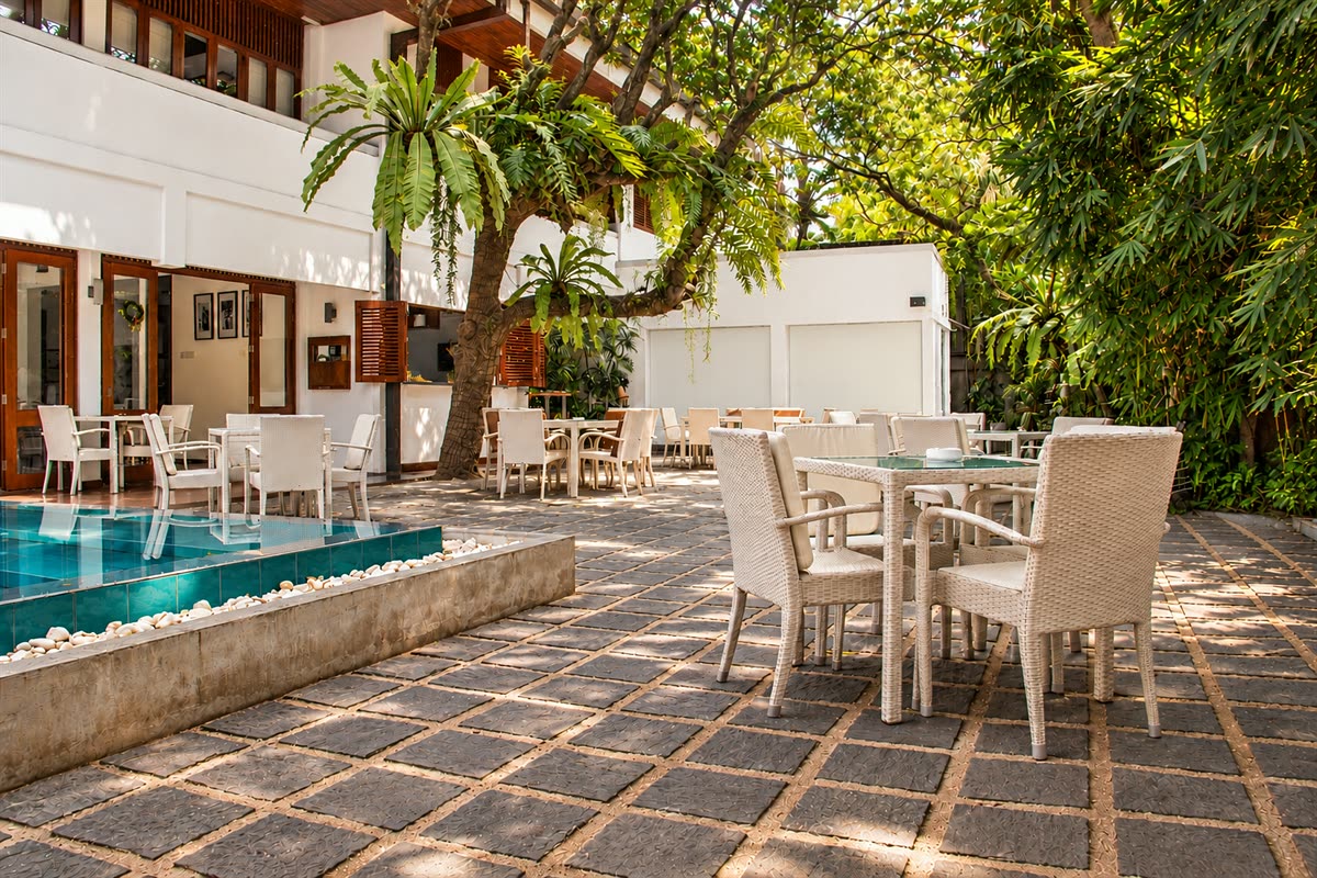Tables set under the old tree beside the pool at Amber Poolside, sunlight through the leaves onto the stone floor.