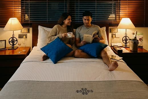 A couple sitting up in bed with cups of tea in a room at Colombo Court, the lamps lit and the blinds down.