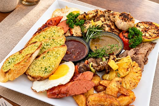 The sharing platter at Amber Poolside: grilled meats, sausages, a fried egg, garlic bread, wedges and a pot of sauce on a long white plate.