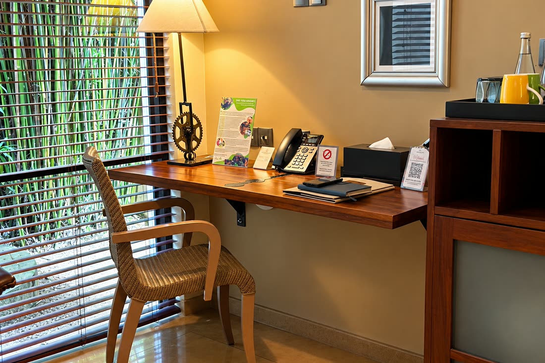 The writing desk in a Deluxe Room, a cane chair and a lamp beside timber blinds.