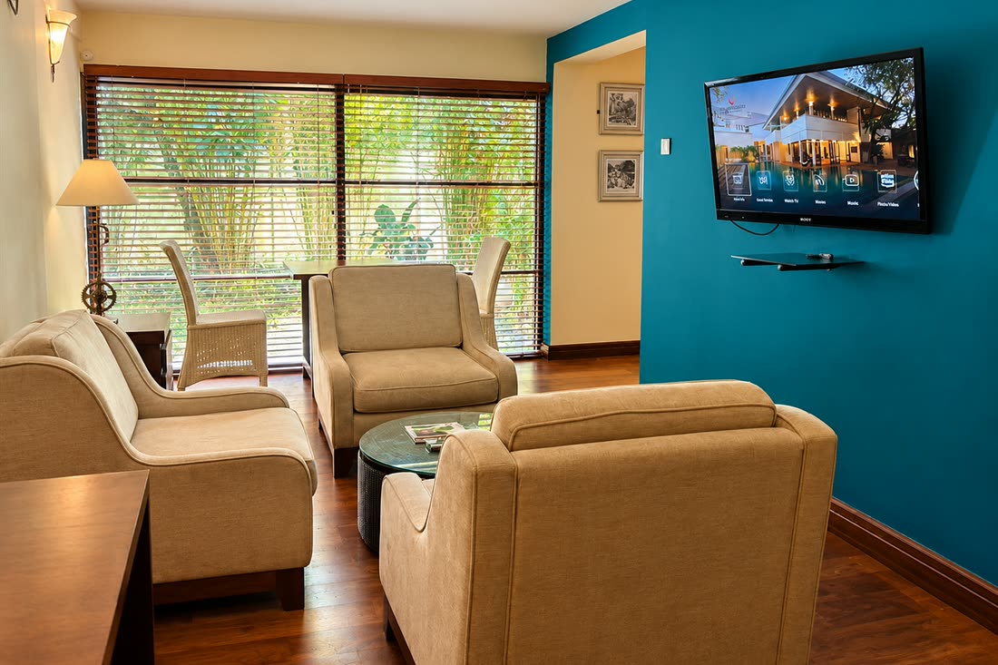 The sitting room in the Luxury Duplex Suite, pale armchairs and a sofa against a teal wall, the television on the wall.