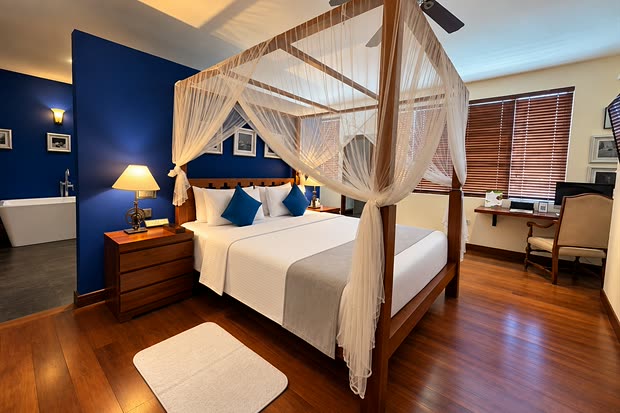 The Luxury Honeymoon Suite at Colombo Court, a four-poster bed under a white net against a deep blue wall, lamps lit either side.