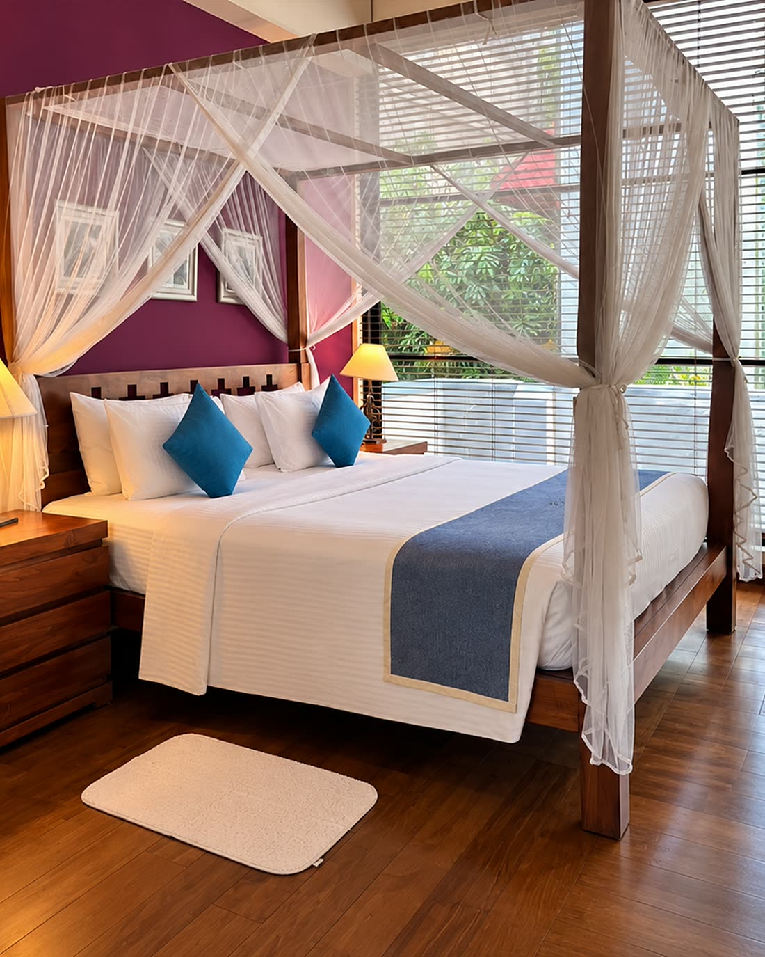 A Luxury Junior Suite with a timber four-poster bed under a white net beside a window of trees.