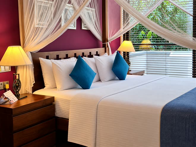The bed in a Luxury Junior Suite at Colombo Court, white linen and blue cushions against a plum wall, lamps lit either side.