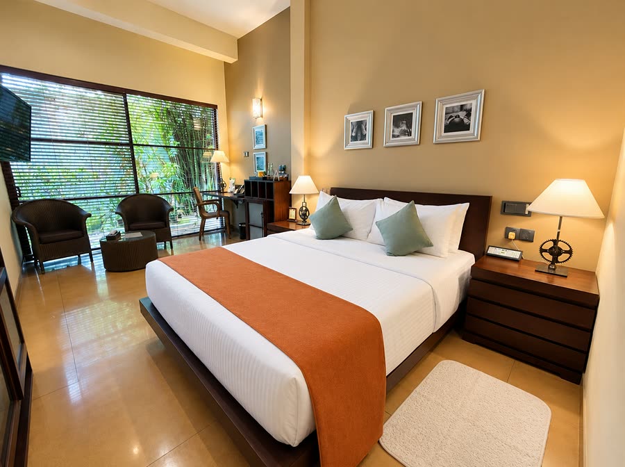 A Deluxe Room at Colombo Court with a queen bed, an orange runner across white linen, and armchairs at a window onto the garden.