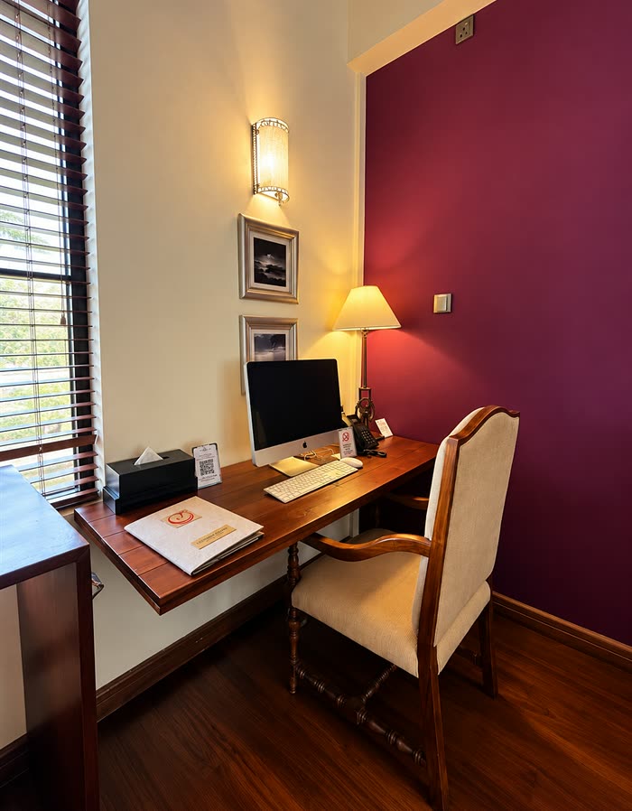 The desk nook in a Luxury Junior Suite at Colombo Court, a lamp lit beside the screen under a plum wall.
