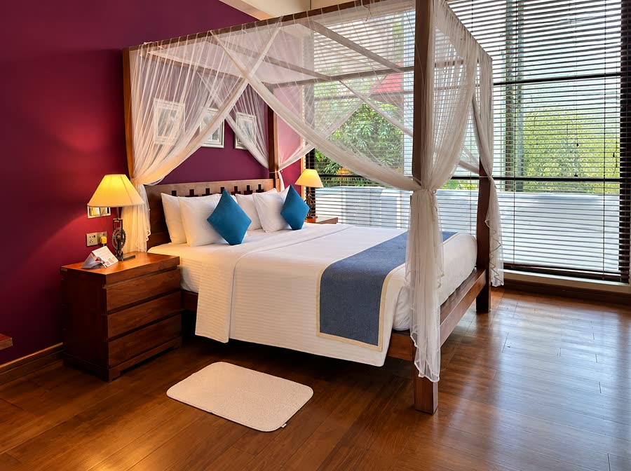 A Luxury Junior Suite at Colombo Court, a four-poster bed under a sheer white net against a plum wall, the window full of trees.
