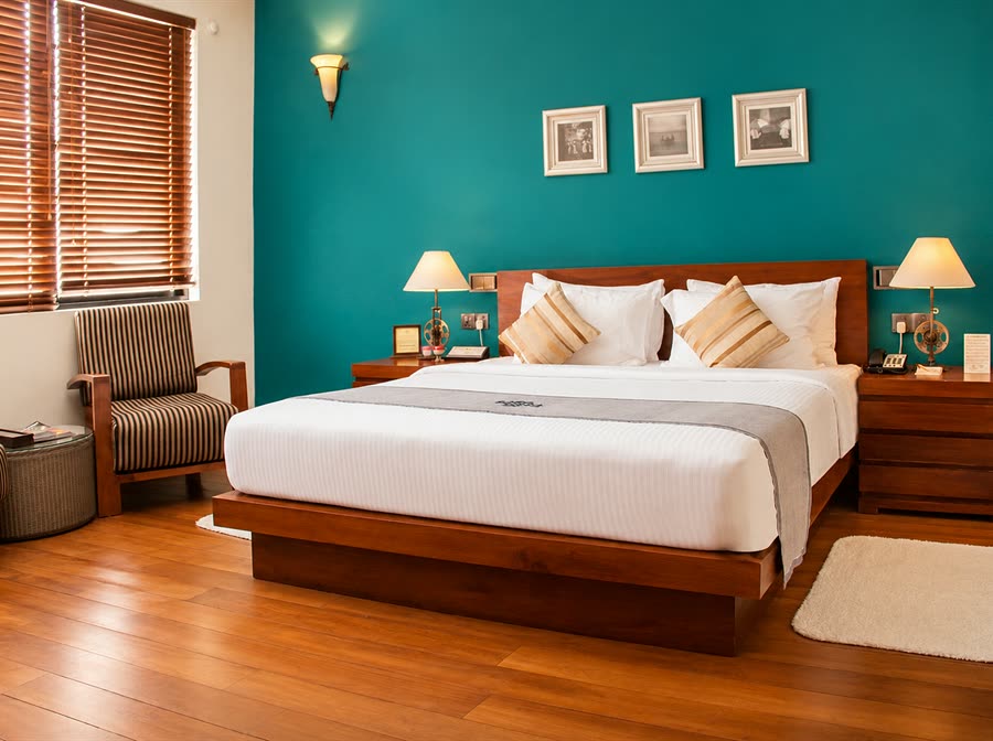 A Superior Deluxe room at Colombo Court, a king bed with cream cushions against a teal wall, framed prints above and timber blinds at the window.