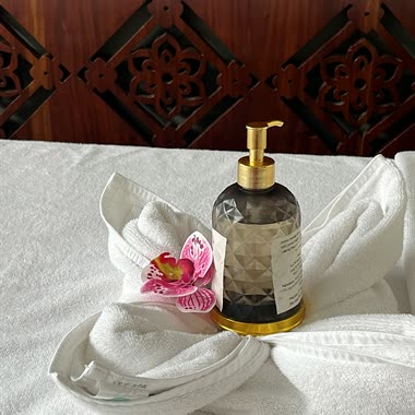 A bottle of massage oil set in a folded towel with a pink flower on a treatment bed.